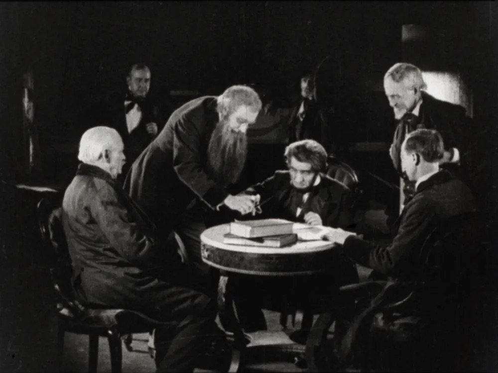 Black and white photo of a group of old men huddles around a table.